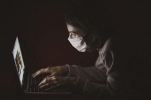 person typing on laptop in the dark with a mask