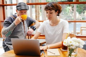 A man drinks coffee and works with a woman who is typing on a computer.