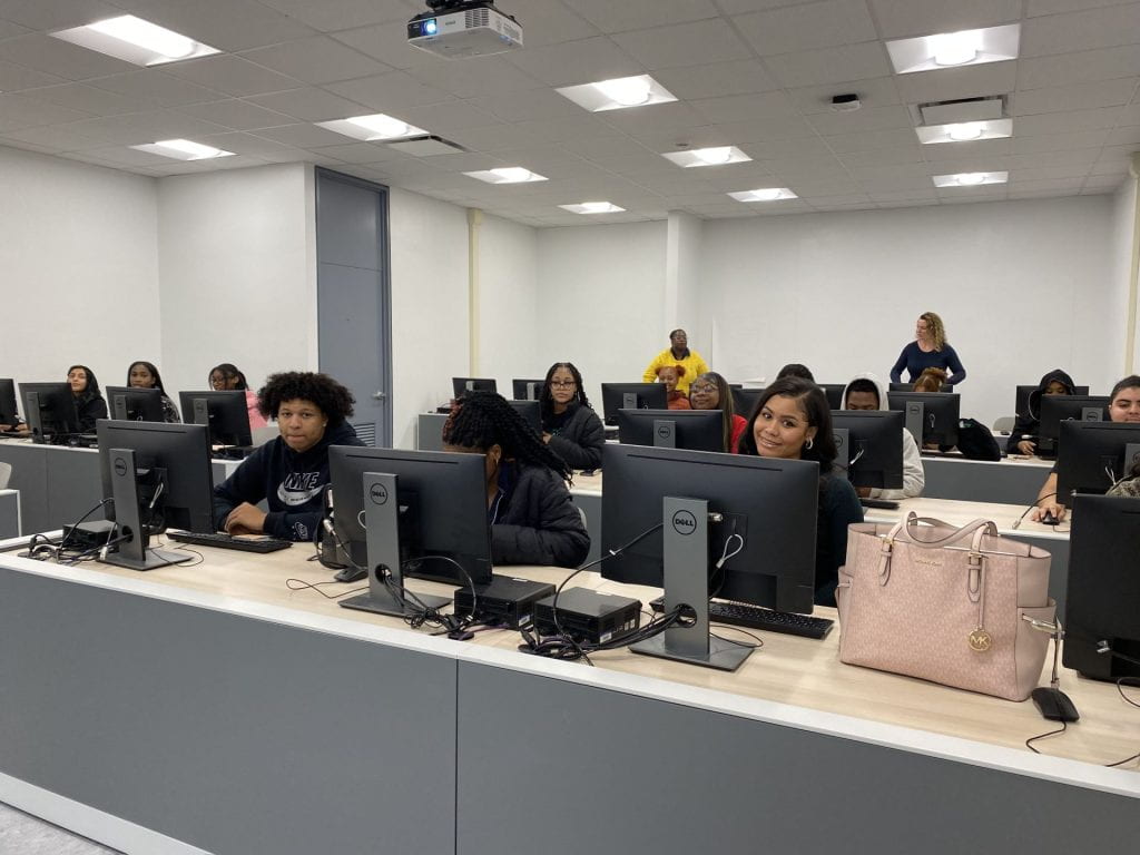 Students sitting at computers, many of them smiling, with two teachers standing in the background.